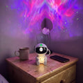 Astronaut Galaxy Star Projector hugging a star on a nightstand by a mug and phone, purple nebula and green stars on the wall