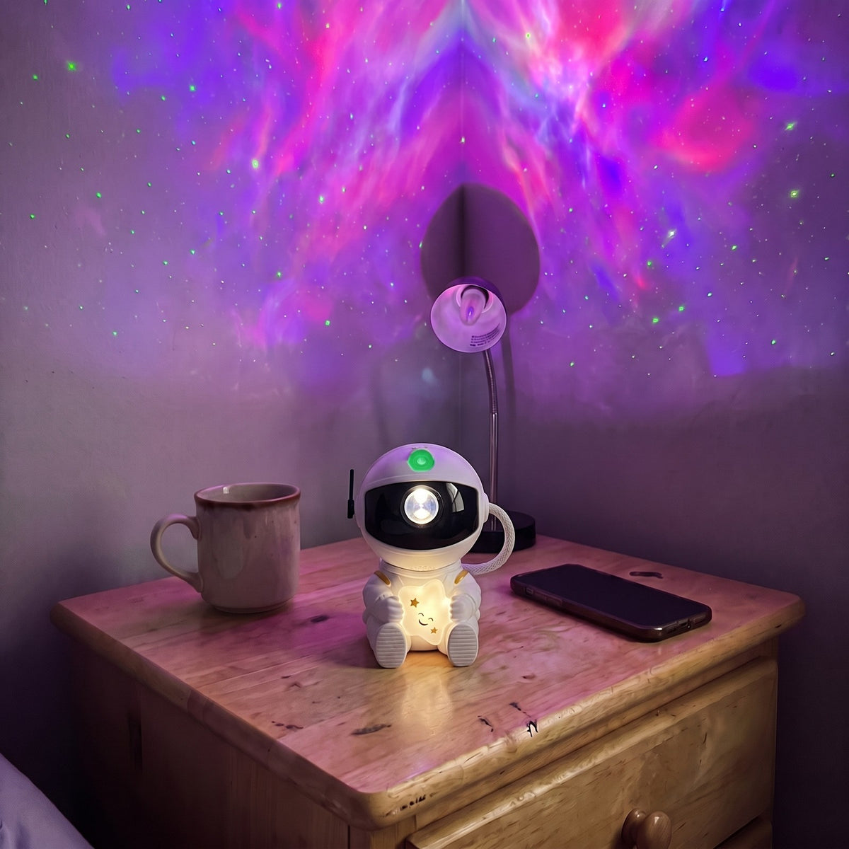 Astronaut Galaxy Star Projector hugging a star on a nightstand by a mug and phone, purple nebula and green stars on the wall