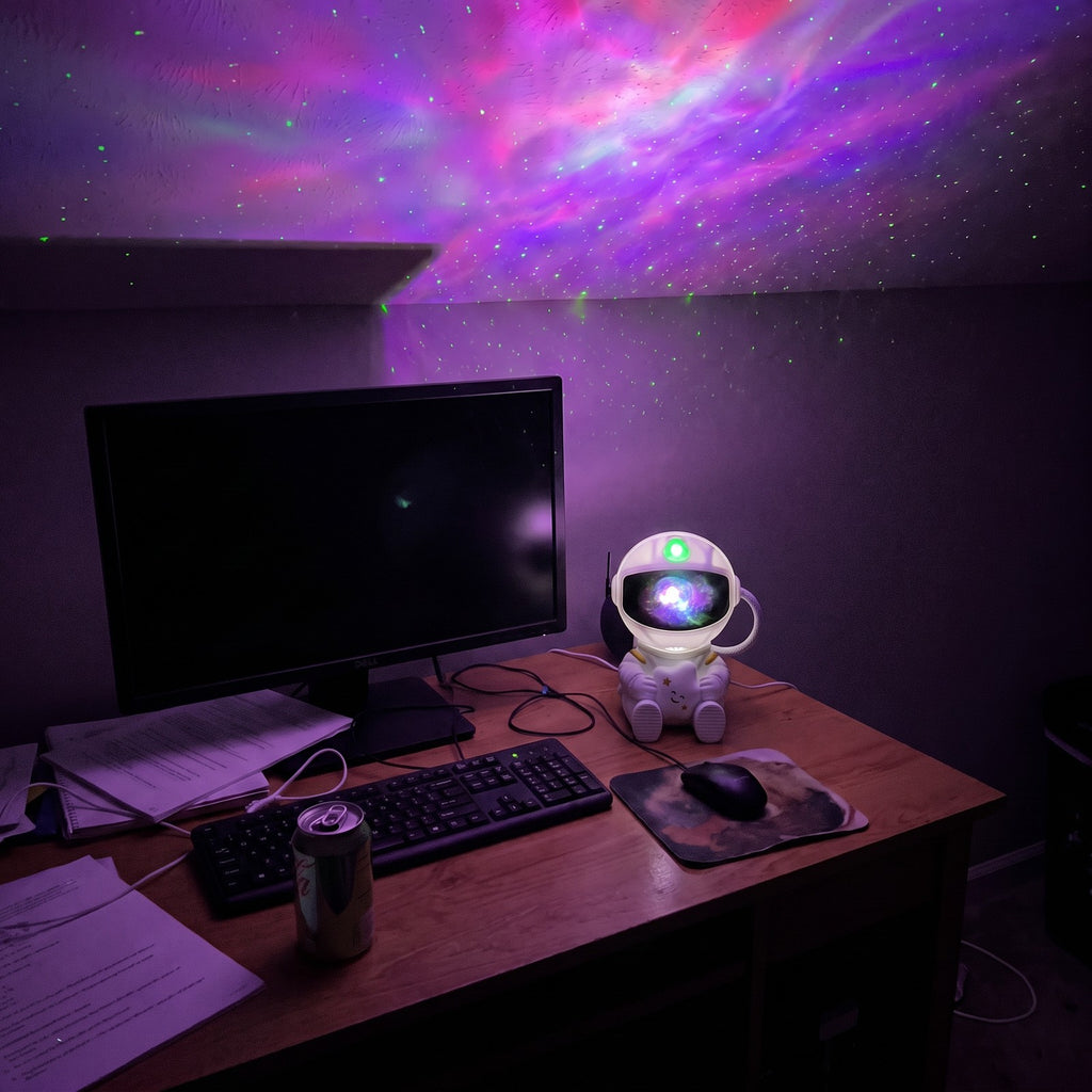 Astronaut projector on a desk beside a monitor, lighting the ceiling with a purple nebula and green stars