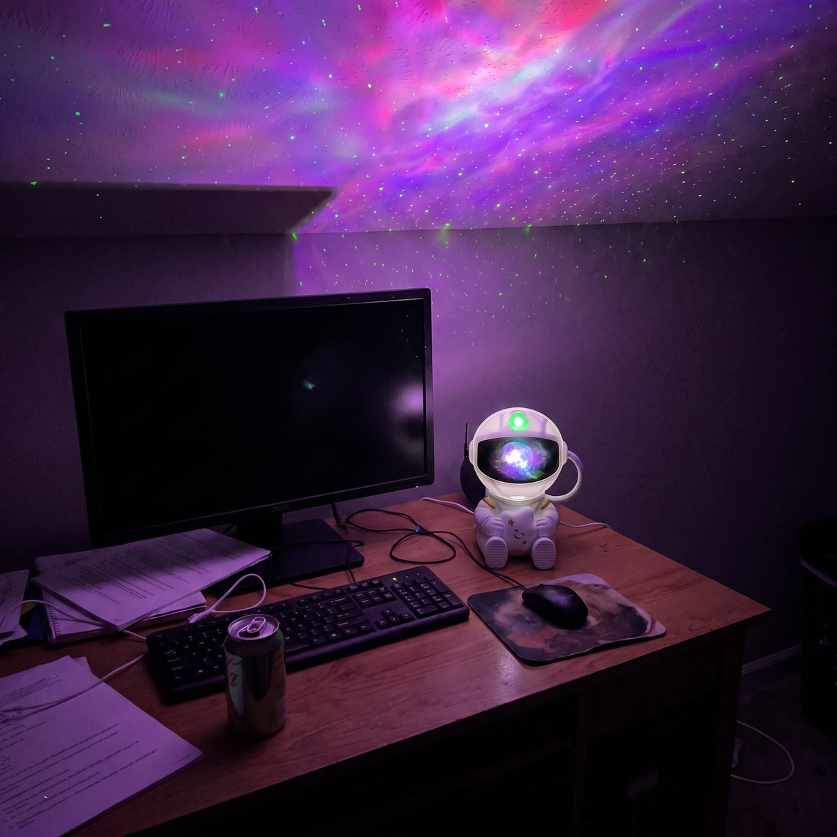 Astronaut projector on a desk beside a monitor, lighting the ceiling with a purple nebula and green stars