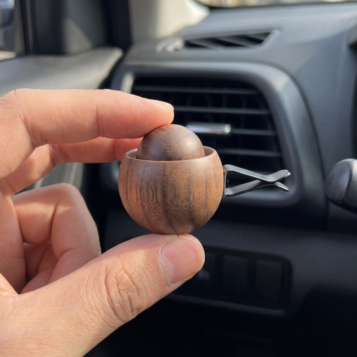The wooden diffuser pinched between two fingertips in front of a dashboard vent, showing its walnut size